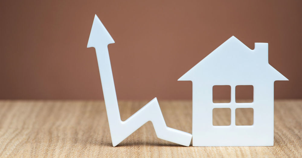 The image depicts a white icon of a house next to a jagged arrow symbol on a wooden surface. The house icon features four square windows, representing a simplified residential structure. The arrow starts with a zigzag pattern, indicating fluctuations, and ends with a sharp upward point, symbolising an increase or growth.